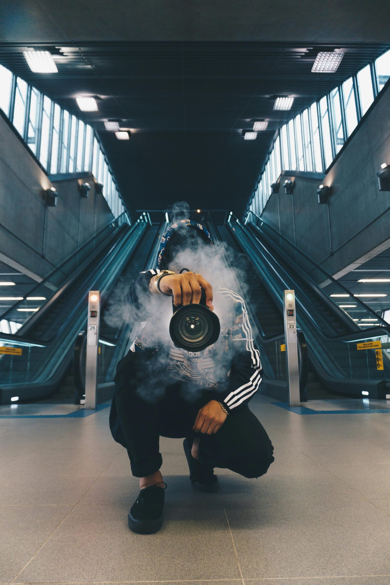 Accueil Person holding a camera in a smoke-filled urban setting with escalators.